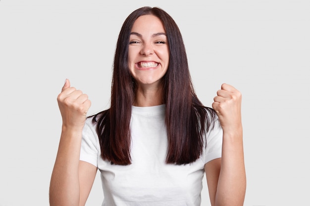 Successful woman clenches fists with happiness, has broad smile, squints face, dressed casual t shirt, isolated over white wall. Carefree brunette girl rejoices success. Victory, determination