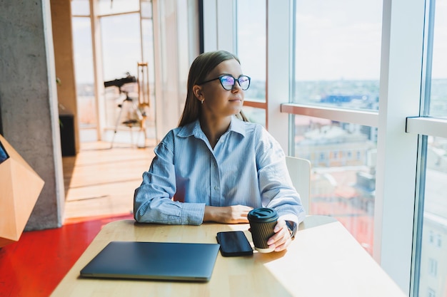 Successful woman is looking at a laptop in a cafe and drinking coffee A young smiling woman in glasses sits at a table near the window with a phone Freelance and remote work Modern female lifestyle