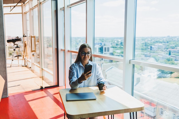Successful woman is looking at a laptop in a cafe and drinking coffee A young smiling woman in glasses sits at a table near the window with a phone Freelance and remote work Modern female lifestyle