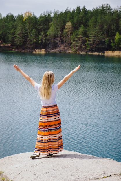 Successful woman traveling near big blue lake, standing on the cliff celebrating victory with her hands up in long skirt