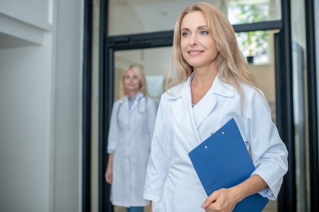 Successful young adult smiling female doctor in uniform with folder walking down corridor and colleague behind