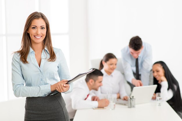 Successful young businesswoman standing proudly in the office and looking at the camera. Her young colleagues working at laptop on desk.