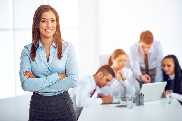 Successful young businesswoman standing proudly in the office and looking at the camera. Her young colleagues working at laptop on desk.