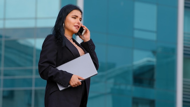 Successful young businesswoman walks down street against background of city building holds laptop