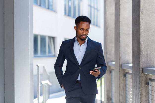 Foto riuscito giovane uomo d'affari afroamericano fiducioso in movimento con il cellulare in mano contro il moderno edificio per uffici sfondo nero centro uomo d'affari cammina utilizzando un telefono cellulare in abito formale