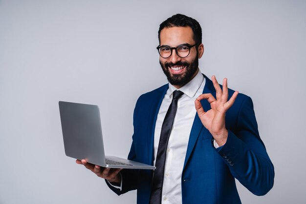 Photo successful young islamic businessman showing okay gesture with laptop isolated over grey background