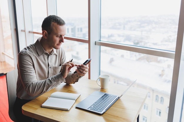 Successful young man is sitting at a table in a caferestaurant indoors working and studying on a laptop computerholding a mobile phone with a blank screen Freelancer mobile office business concept