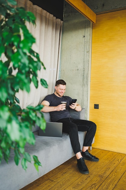 Successful young man in tshirt sitting on sofa with phone and drinking coffee working remotely during startup as a freelancer looking at laptop and smiling