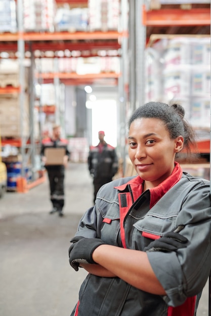 Photo successful young mixed-race female worker of large warehouse crossing arms by chest while standing by rack in aisle against male colleagues