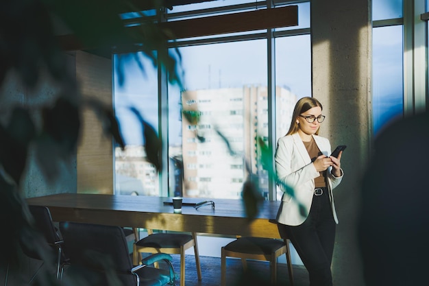 A successful young woman in glasses and a jacket is standing in the office and happily talking on the phone A young manager works in the office and manages the business remotely