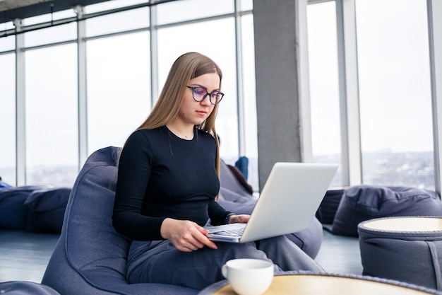 성공한 젊은 여성 매니저는 탁 트인 창가에 있는 부드러운 안락의자에 노트북을 놓고 커피를 마십니다. 새로운 프로젝트에서 일하는 비즈니스 우먼