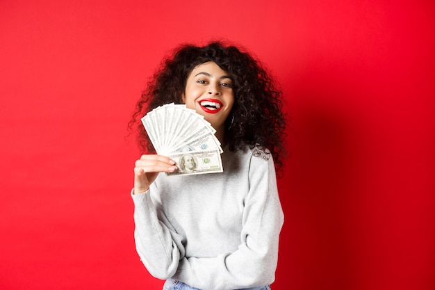 Successful young woman waiving dollar bills and smiling pleased. Rich girl showing money, standing over red wall.