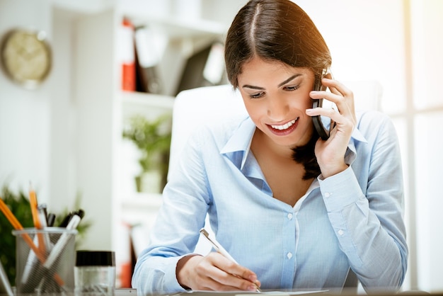 Successful young woman working at her desk in the office. She is using smart phone and writing business report.