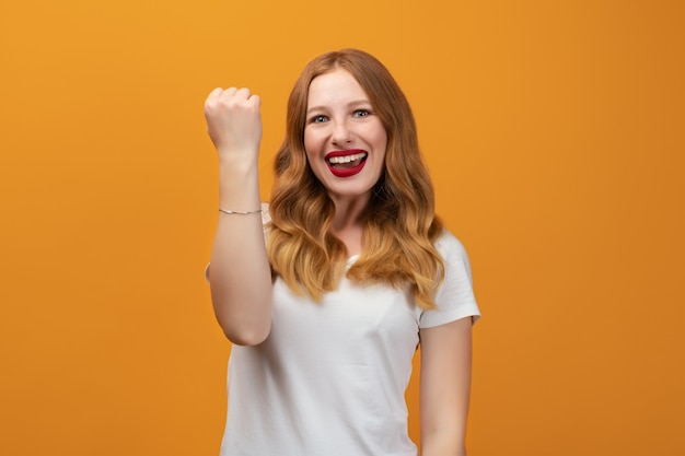 Foto succesvol meisje met lang golvend blond haar, gekleed in een wit t-shirt glimlachend gelukkig, geweldig nieuws vieren, winnaar gebaar maken, staande gele achtergrond