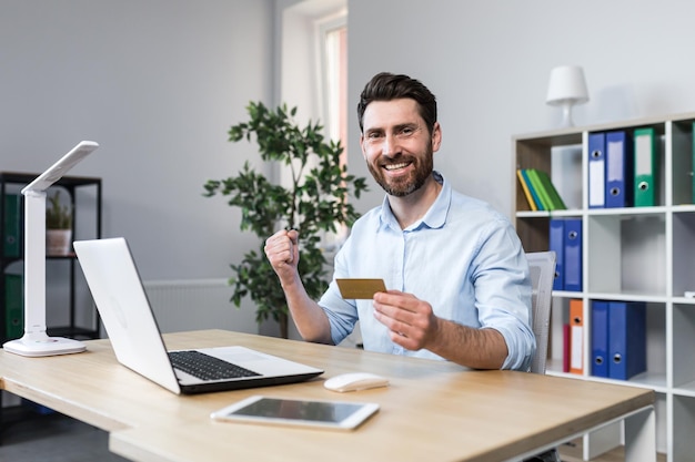 Succesvol online winkelen Succesvolle deal Tevreden jonge man met een creditcard die zijn hand laat zien Ja Zittend aan een bureau op kantoor op de computer