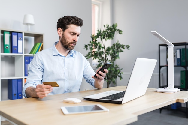 Succesvol online winkelen via een smartphone Succesvolle deal Tevreden jonge man heeft een creditcard en een mobiele telefoon maakt een bestelling Zittend aan een bureau op kantoor op een laptop