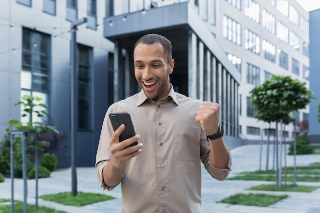 Succesvolle afro-amerikaanse zakenman buiten kantoorgebouw met behulp van smartphone glimlachend en gelukkig