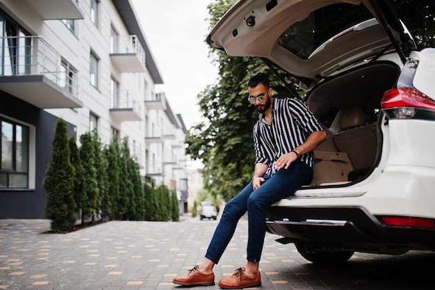 Succesvolle arabische man draagt een gestreept shirt en een zonnebril in de kofferbak van zijn witte suv-auto. Stijlvolle Arabische mannen in transport.