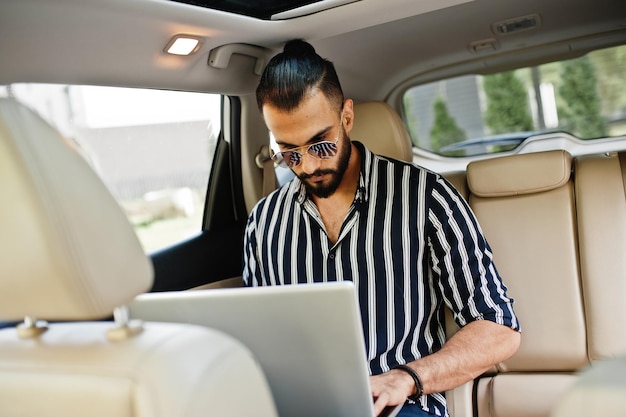 Succesvolle arabische man draagt een gestreept shirt en zonnebril pose in witte suv-auto met laptop in handen.