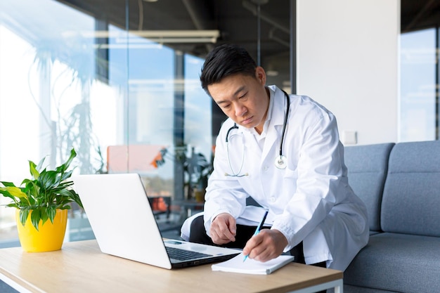 Succesvolle Aziatische arts die aan laptop in een moderne kliniek werkt.