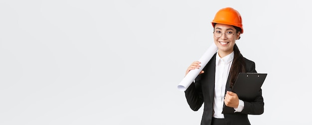 Succesvolle Aziatische vrouwelijke architect in pak en helm met blauwdrukken en klembord met notities inspecteur kijken naar bouwwerkzaamheden glimlachend op camera witte achtergrond