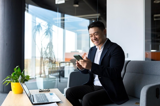 Succesvolle Aziatische zakenman viert overwinning, kijkt blij naar de telefoon en verheugt zich over het zitten in een modern kantoor