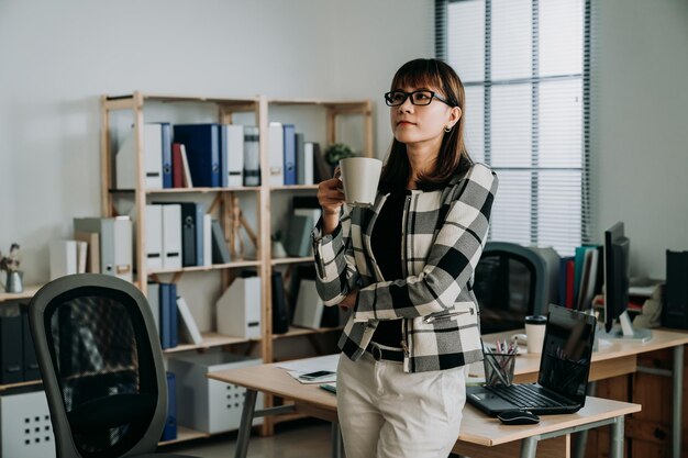 succesvolle Aziatische zakenvrouw die tegen het bureau staat, staart in de verte in contemplatie terwijl ze 's ochtends op haar kantoor koffie drinkt.