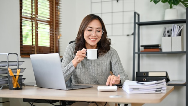 Succesvolle Aziatische zakenvrouw of accountant die rekenmachine gebruikt terwijl ze koffie drinkt aan het bureau