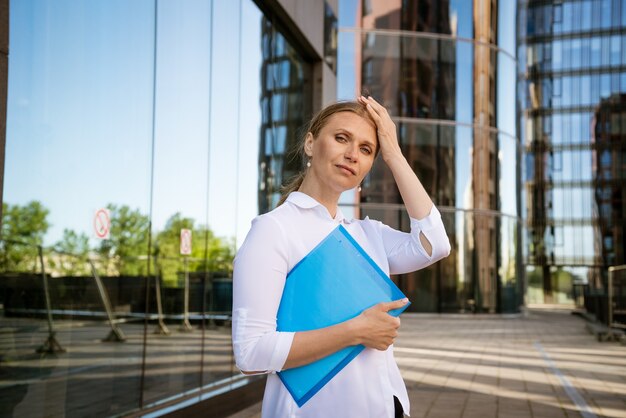 Succesvolle bedrijfsvrouw in wit overhemd met blauwe map voor papieren staat op kantoorgebouw