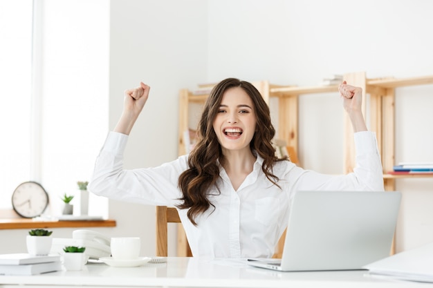 Succesvolle bedrijfsvrouw met wapens die omhoog in modern bureau zitten.