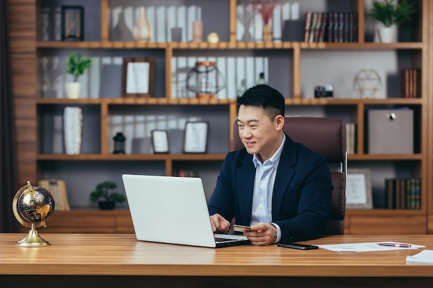 Succesvolle en gelukkige zakenman die online winkelt in een online winkel Aziatische man aan het werk in een modern kantoor met een laptop met een creditcard