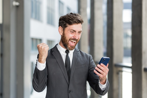 Succesvolle en gelukkige zakenman schreeuwt met vreugde en voldoening om te winnen, leest goed nieuws van een mobiele telefoon, buiten kantoor kijkt naar de camera