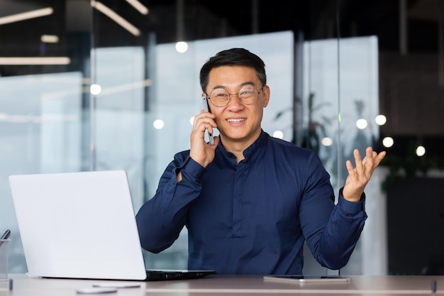 Succesvolle en vrolijke aziatische zakenman die aan de telefoon praat met klanten binnen het volwassen kantoor