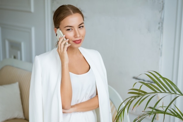 Succesvolle jonge en mooie vrouw in een witte jas en jurk praten aan de telefoon