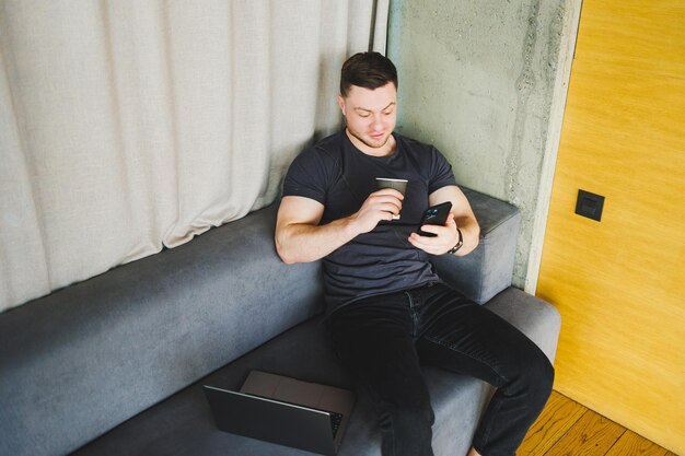 Succesvolle jonge man in t-shirt zittend op de bank met telefoon en koffie drinken op afstand werkend tijdens opstarten als freelancer die naar laptop kijkt en glimlacht