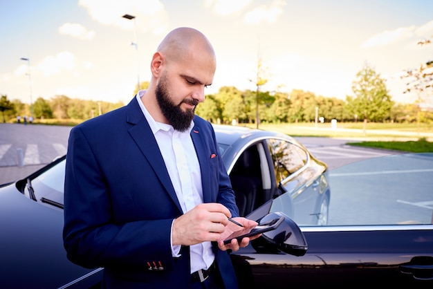 Succesvolle jonge man met een telefoon in de buurt van een auto op een parkeerplaats.