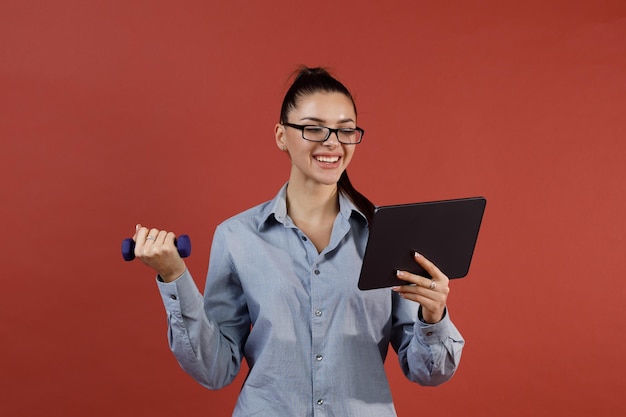 Succesvolle jonge moderne zakenvrouw in blauw shirt warming-up met halter op kantoor tijdens het werken aan tablet pad Multitasking en time management concept