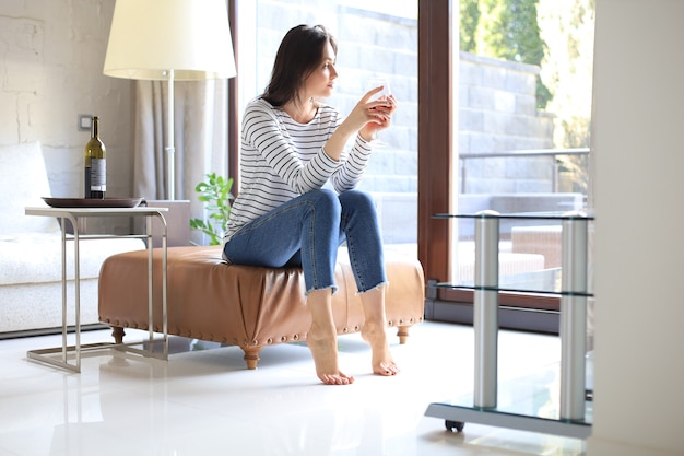 Succesvolle jonge mooie vrouw zittend op een fauteuil in de woonkamer, rode wijn drinken.