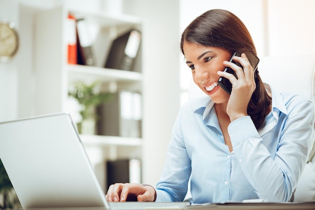 Succesvolle jonge vrouw die aan haar bureau op kantoor werkt. Ze gebruikt slimme telefoon en werkt op laptop.