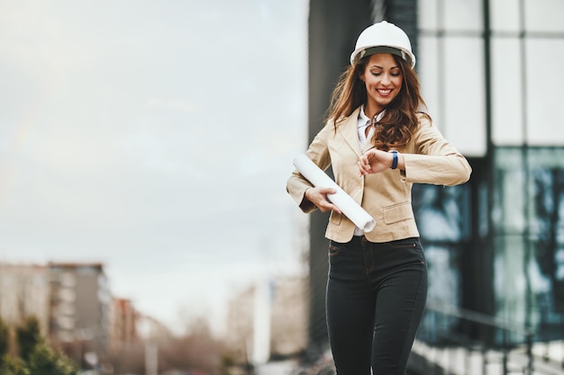 Succesvolle jonge vrouwelijke ingenieur met witte helm en blauwdrukken in haar handen voor kantoordistrict.