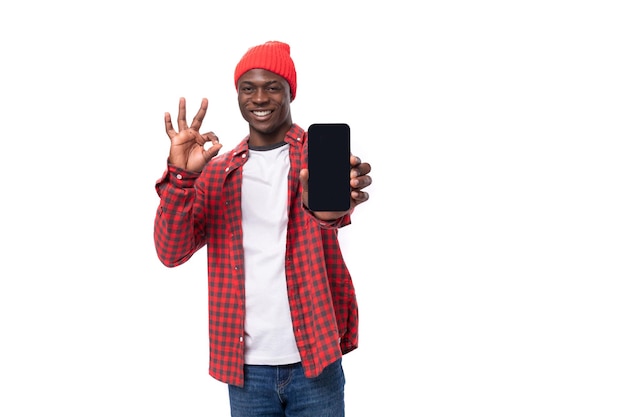 Succesvolle knappe donkere Amerikaanse man gekleed in rood shirt en pet toont smartphone