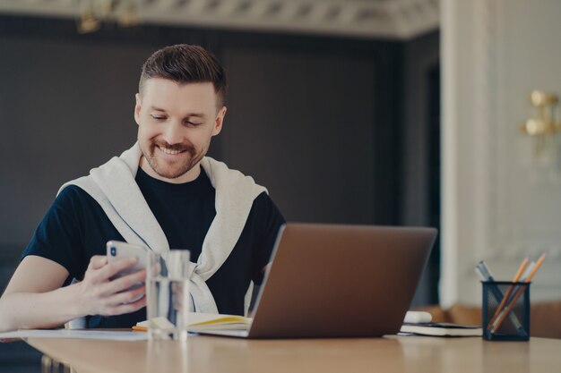 Succesvolle knappe mannelijke freelancer in vrijetijdskleding kijkend naar smartphone en glimlachen tijdens het lezen van berichten of goed nieuws, zittend op zijn werkplek voor laptop thuis. Bedrijfs- en werkconcept