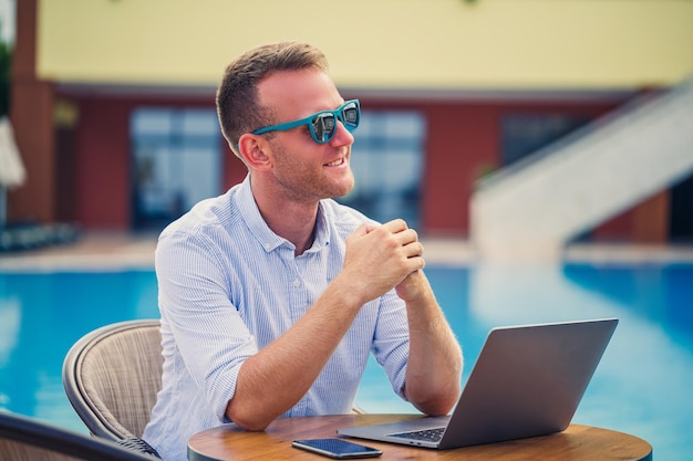 Succesvolle knappe mannelijke zakenman in zonnebril werkt op een laptop bij het zwembad Werken op afstand Freelancer