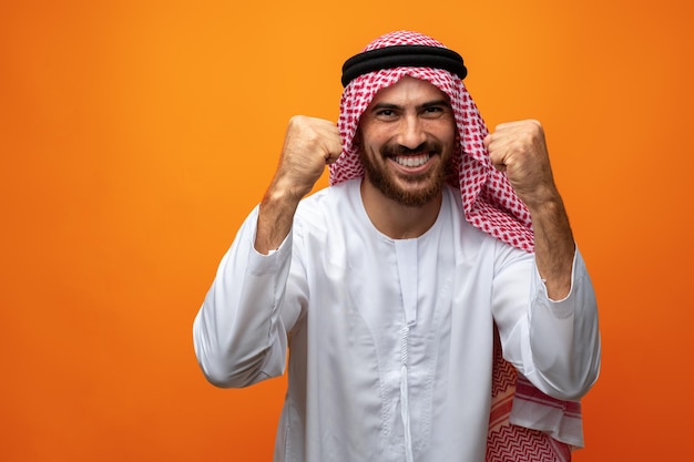 Succesvolle lachende jonge Arabische man viert overwinning op oranje achtergrond