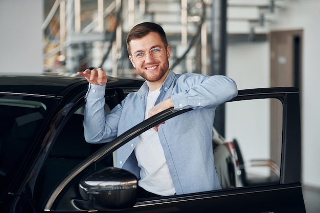 Succesvolle man met een bril die binnenshuis in de buurt van een gloednieuwe auto staat