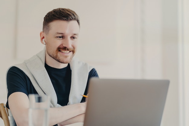 Succesvolle mannelijke freelancer met webconferentie met collega's die oortelefoons en laptop gebruiken, blij zijn om goed nieuws te horen, vrolijke zakenman in vrijetijdskleding die via videogesprek op kantoor praat