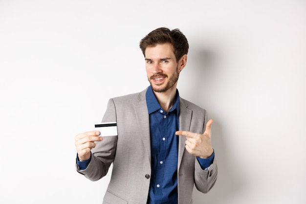 Succesvolle mannelijke ondernemer wijzend op plastic creditcard en glimlachen, bank aanbevelen, staande op een witte achtergrond.