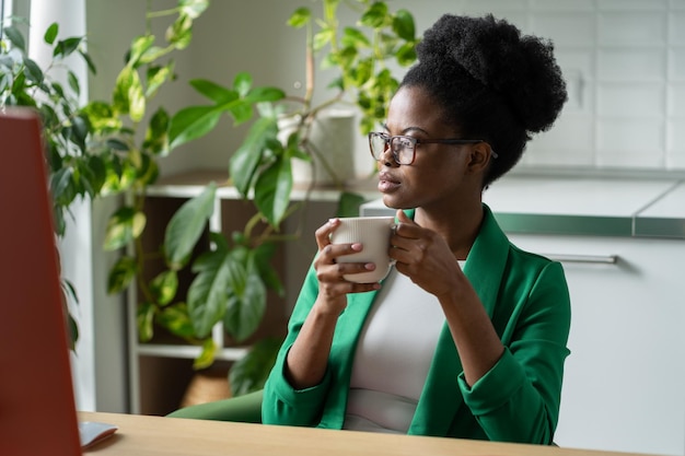 Succesvolle peinzende afro-amerikaanse vrouw die koffie drinkt, zit aan tafel met computer in thuiskantoor