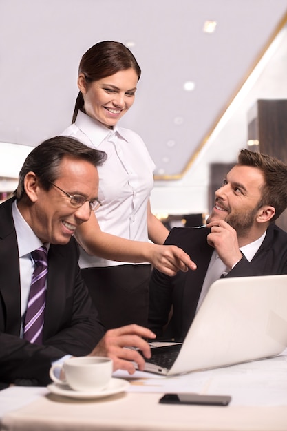 Succesvolle ploeg. Twee vrolijke zakenmensen in formele kleding die aan tafel zitten en glimlachen terwijl een vrouw in een wit overhemd een computermonitor aanwijst