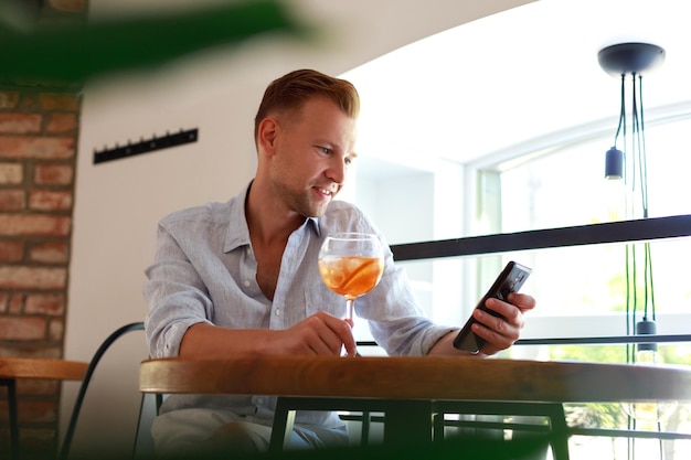 Succesvolle rijke zakenman in hemd die cocktail drinkt en smartphone vasthoudt aan tafel in restaurant